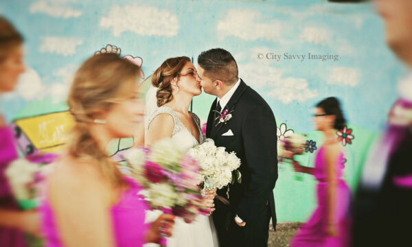 Fernando and Mollie share a kiss in front of a colorful mural, while bridesmaids in pink dresses stroll by, holding bouquets.