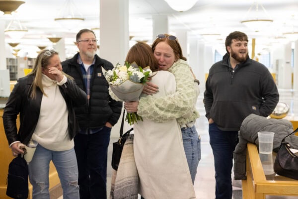 Emotional hug as friends and family celebrate a surprise proposal, with bouquet in hand and joyful reactions all around