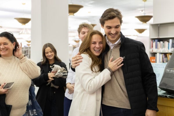 Smiling fiance embracing as friends gather behind them in a library, celebrating a joyful surprise proposal moment.