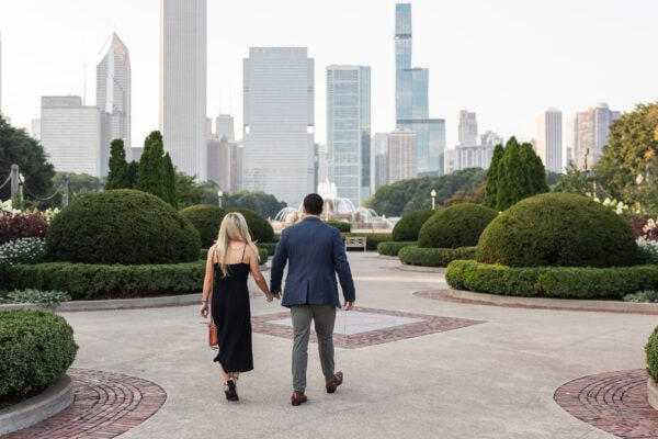 Couple walking hand in hand toward Buckingham Fountain with Chicago skyline in the background before their surprise proposal