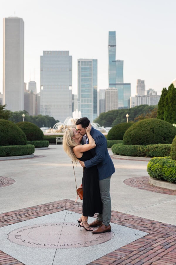 Couple kissing on a plaza near Buckingham Fountain with the Chicago skyline behind them after a romantic surprise proposal.