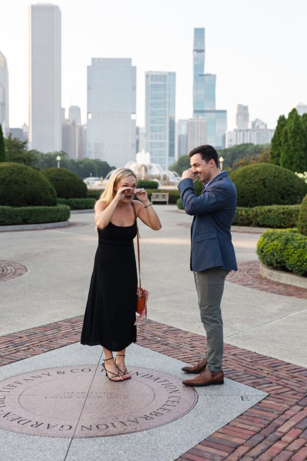 Couple wiping tears of joy after a surprise proposal near Buckingham Fountain with the Chicago skyline behind them.