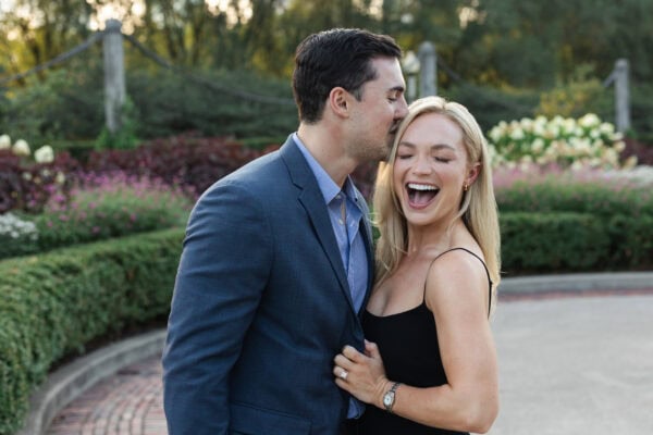 Man kisses partner’s cheek as she laughs, celebrating their engagement in a garden after a surprise proposal