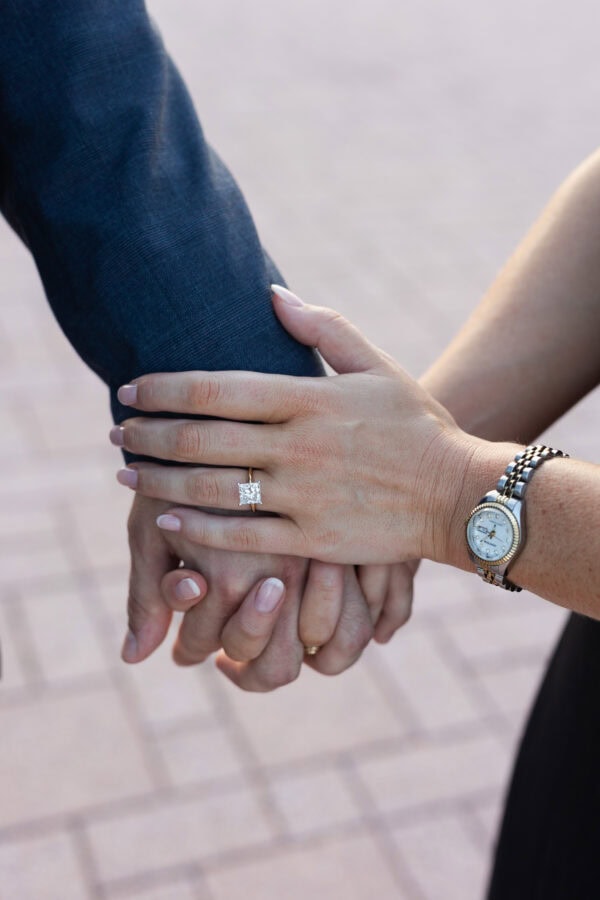 Close-up of hands holding with a diamond engagement ring and watch visible, capturing an intimate moment after a surprise proposal