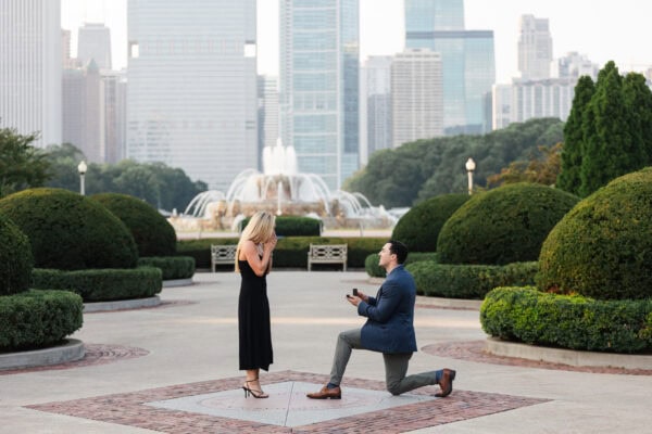 Man kneels to propose to surprised partner near Buckingham Fountain with Chicago skyline in the background.