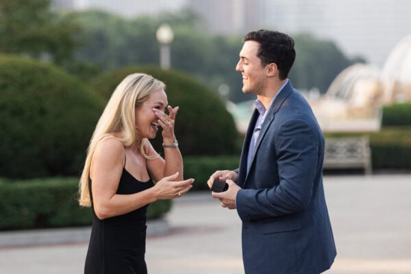Woman reacts with joyful surprise as partner holds ring box during a proposal in a garden with the chicago skyline in the background.
