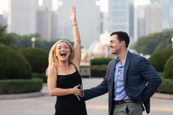 Woman celebrates in excitement after a surprise proposal as her partner smiles, with Chicago skyline in the background.