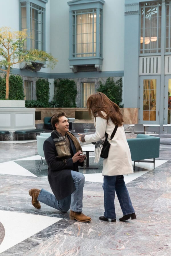 Man kneeling and proposing with a ring to a surprised woman inside the bright, glass-ceiling atrium of the Winter Garden at Harold Washington Library in Chicago, with marble floors, tall windows, and soft natural light.
