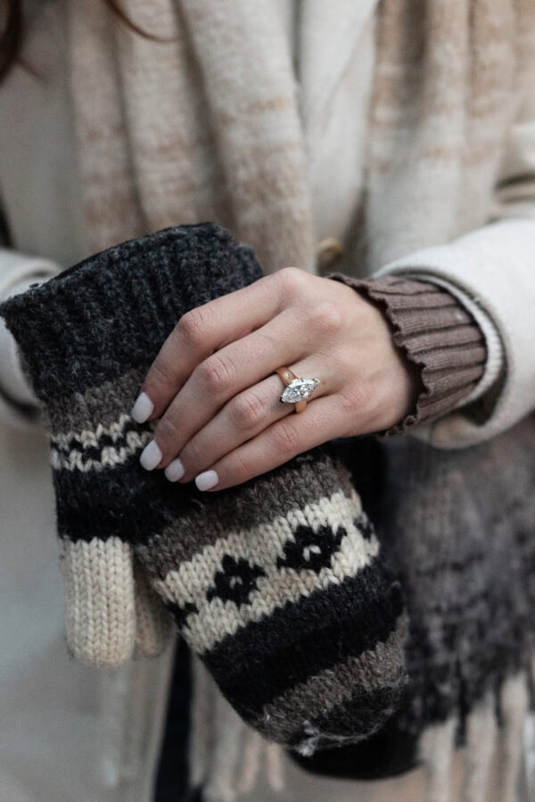 Close-up of a hand wearing an oval diamond engagement ring, holding a knit mitten, capturing a cozy surprise proposal detail.