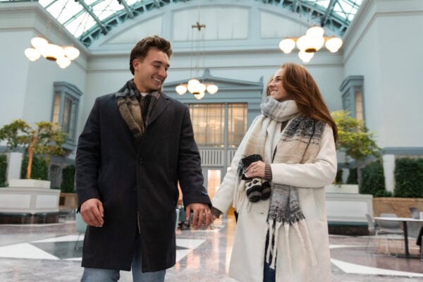 Couple holding hands and smiling while walking through a bright indoor atrium, celebrating a joyful surprise proposal moment.