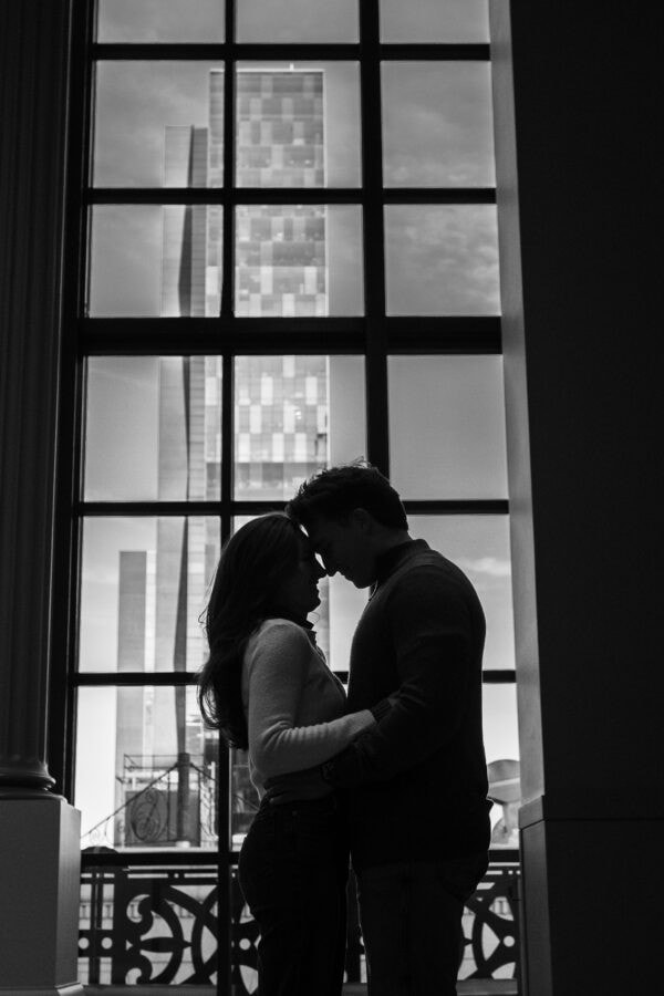 Silhouetted couple kissing by large windows with city skyline at sunset, capturing a romantic moment after a surprise proposal.