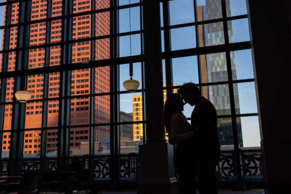 Silhouetted couple kissing by large windows with city skyline at sunset, capturing a romantic moment after a surprise proposal.