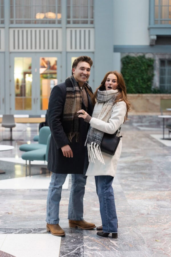 Couple smiling after surprise proposal in bright indoor atrium, woman showing engagement ring with joyful, surprised expression