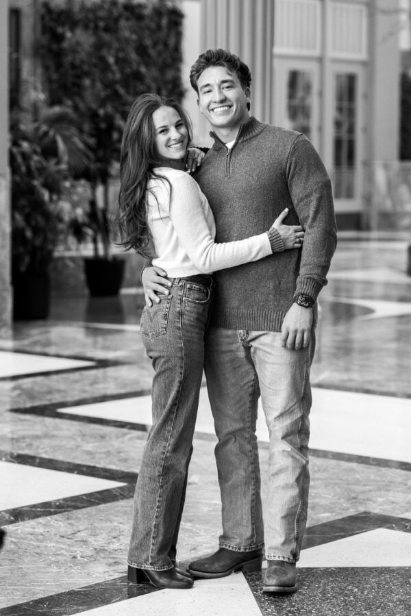 Black-and-white portrait of a smiling couple embracing in a bright atrium after a joyful surprise proposal.