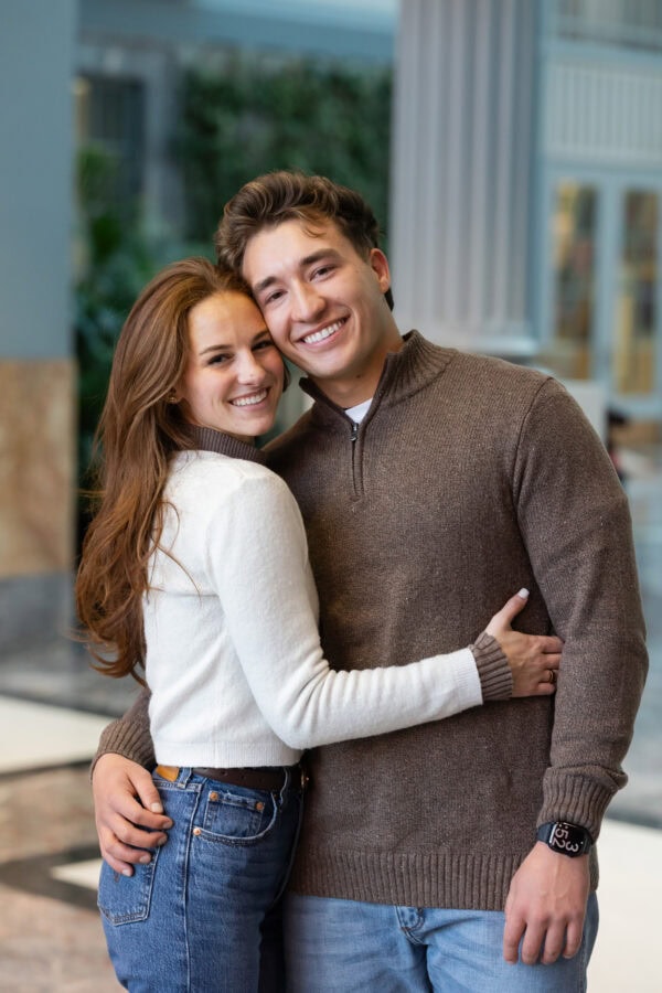 Smiling couple poses for a photo in a bright atrium, celebrating their engagement after a joyful surprise proposal.