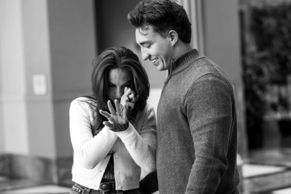 Black-and-white image of a woman wiping tears of joy looking her engagement ring as her partner smiles during a surprise proposal.