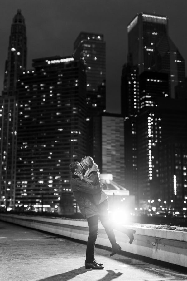 Couple embraces at night along Chicago's lakefront with glowing skyline lights during a romantic engagement photo session
