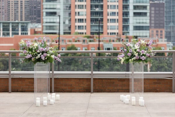 Purple and white floral arrangements are sitting on top of acrylic columns with white candles at the base which is all set up in front of a view of chicago buildings downtown.