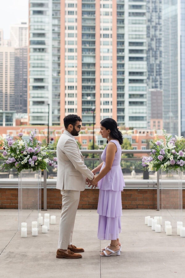 a man and woman hold hands while looking into each others eyes before he proposes to her. They stand in front of floral arrangements.