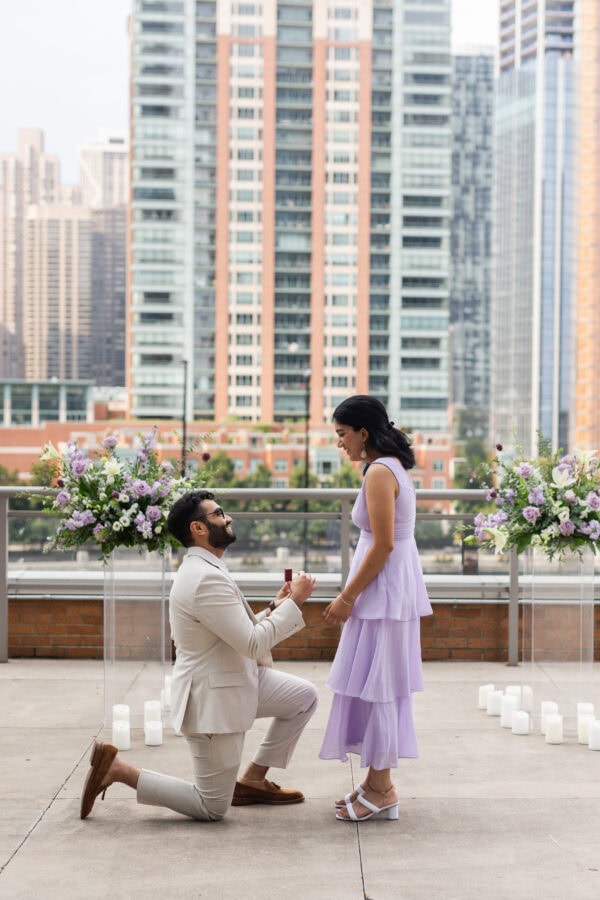 a man in a tan suit is on one knee proposing to his fiance dressed in purple standing in front of purple floral arrangements and city buildings