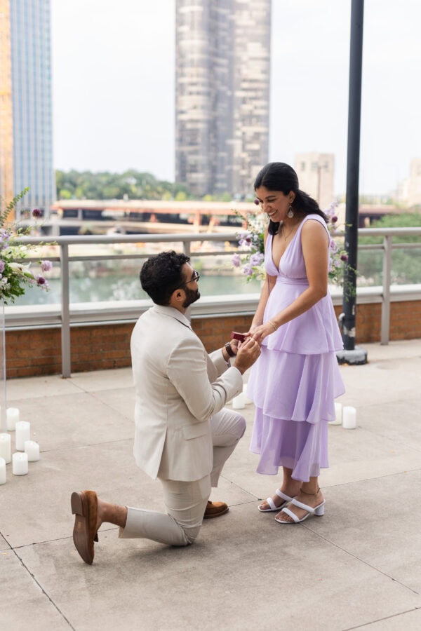 a man in a tan suit is on one knee proposing to his fiance dressed in purple standing in front of purple floral arrangements and city buildings