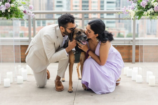 a well dressed newly engaged couple gives their dog in a tuxedo a kiss