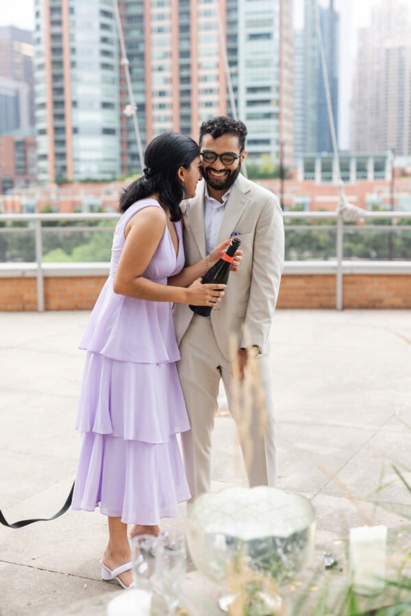 a happy engaged couple laughs while they open a bottle of champagne