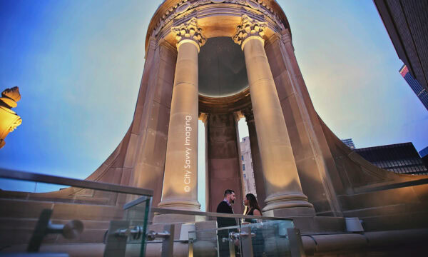 Luis and Michelle stand under a grand, circular stone structure with tall columns, as the LondonHouse rooftop sets a magical backdrop against the clear sky at dusk for Luis's surprise proposal.