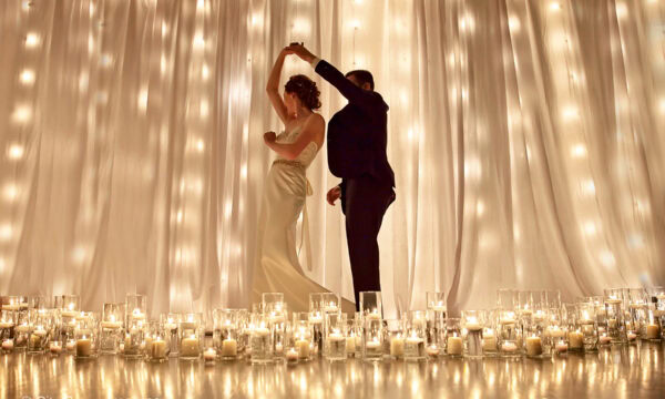 A couple dances in formal attire surrounded by glowing candles in glass holders, with white draped curtains and string lights in the background, creating a romantic wedding scene.