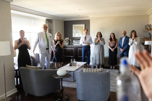 a group of 8 people stand in a hotel room yelling surprise at a couple entering the room