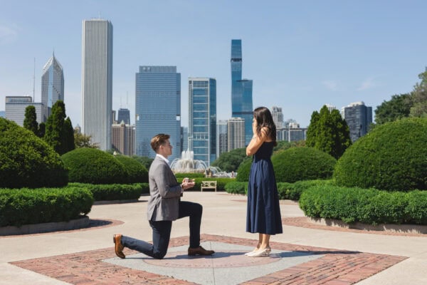 a man is down on one knee proposing to his girlfriend in front of the chicago skyline on a summer day