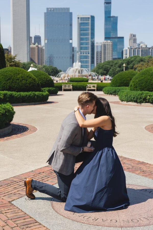 a man is down on one knee proposing to his girlfriend and sharing a kiss in front of the chicago skyline on a summer day