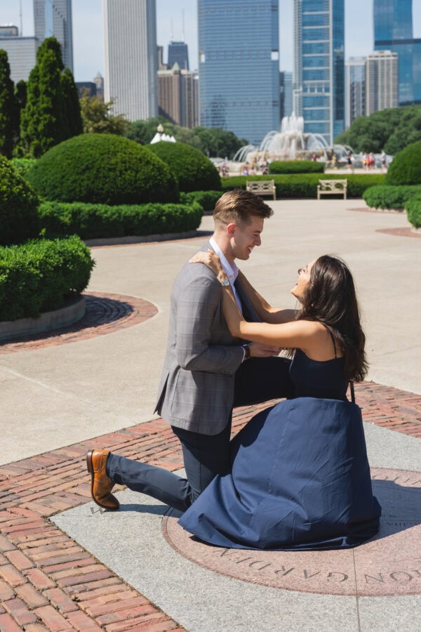 a man is down on one knee proposing to his girlfriend in front of the chicago skyline on a summer day