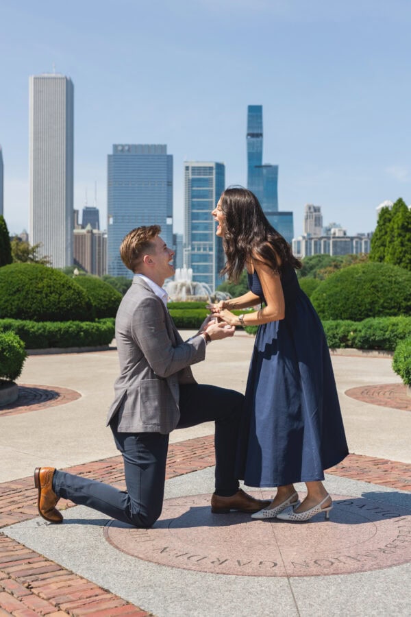 a man is down on one knee proposing to his girlfriend in front of the chicago skyline on a summer day
