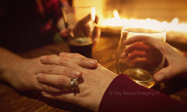 Hands resting on a wooden table, one adorned with an engagement ring, capture the intimacy of the moment. Nearby, drinks and a lit fireplace add warmth to this Chicago engagement session scene.