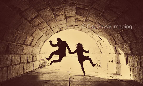 In a Chicago engagement session, silhouettes of two people joyfully jumping in a stone tunnel are backlit by a radiant glow.