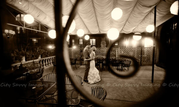 A couple embraces on Salvatores' charming outdoor terrace in Chicago, under round hanging lights and a draped ceiling, surrounded by empty metal chairs and tables, evoking an intimate wedding atmosphere.