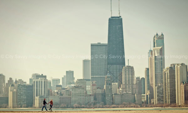 Two people walk hand in hand on the sandy shores of North Avenue Beach, with the Chicago skyline painting a picturesque backdrop on their unforgettable engagement day.