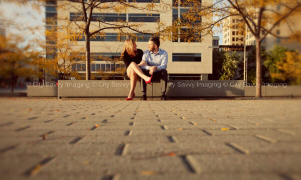 Nick and Morgan sit on a bench in an urban park, surrounded by autumn trees and city buildings. It's the perfect setting for their engagement session.