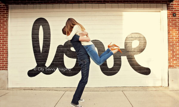 Paul & Katherine share a heartfelt embrace in front of a Chicago garage door, where the word "love" is painted in large, stylized letters—a fitting backdrop for their surprise proposal.
