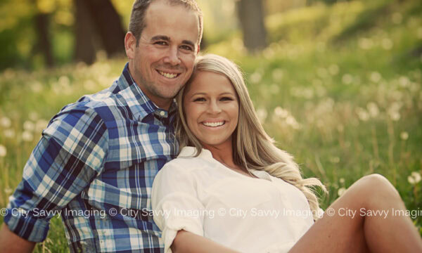 Ashley and Adam are sitting on the grass in a park, celebrating their engagement. The man, Adam, wears a plaid shirt and jeans, while Ashley looks lovely in her white top. They're smiling with joy, surrounded by trees in the background.