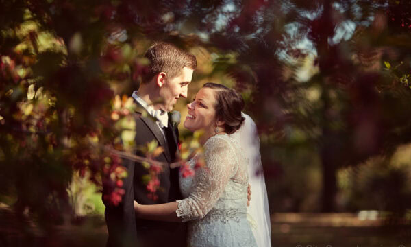 Annie & Dan stand close in an outdoor setting at Abbey Station Wedding, surrounded by blurred greenery and pink flowers, smiling at each other.