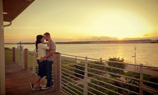 Elena & Dustin's engagement session unfolds as they stand on a wooden deck by the river, embracing and gazing at each other during a breathtaking sunset.