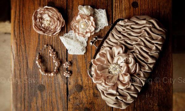 A ruched satin clutch with floral embellishment is displayed alongside matching floral pins, a pearl bracelet, and earrings on a wooden surface, perfect for adding elegance to any Quad Cities wedding.