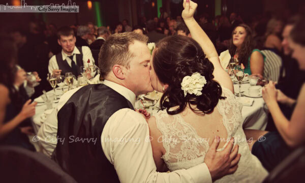 At the Arboretum Club Wedding, a couple shares a kiss at the lively reception, surrounded by seated guests. The bride, adorned with a floral hairpiece, raises her right arm joyfully.