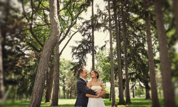 Mike & Sara, dressed in elegant formal attire, embrace and smile among tall trees in a sunlit park setting, capturing the warmth of their brunch wedding.