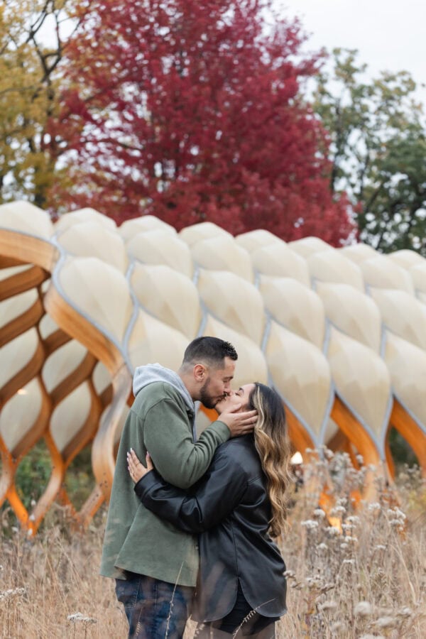a couple shares a kiss in front of Chicago's Peoples Gas Pavilion surrounded by fall trees