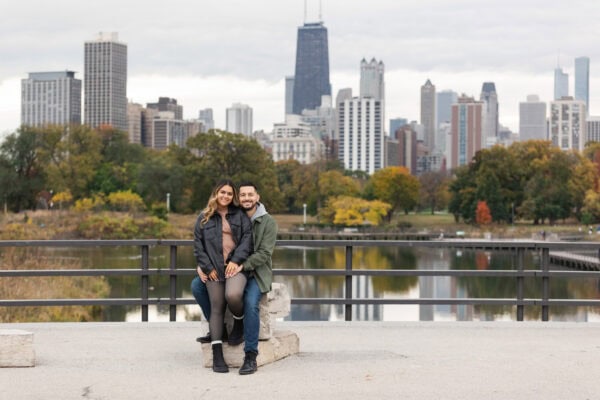 A newly engaged couple poses for a photo sitting on a rock in front of the chicago skyline in Lincoln Park