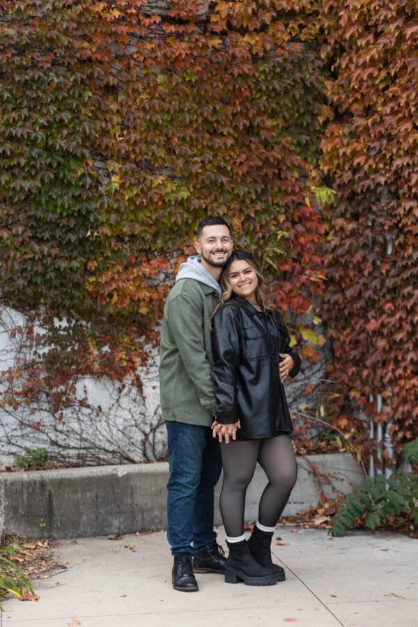 A newly engaged couple poses for a photo in front of a colorful wall of ivy