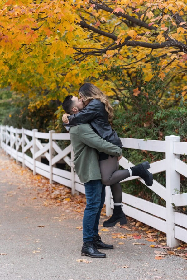 a couple shares a kiss in front of colorful fall trees and white fence in Chicagos Lincoln Park